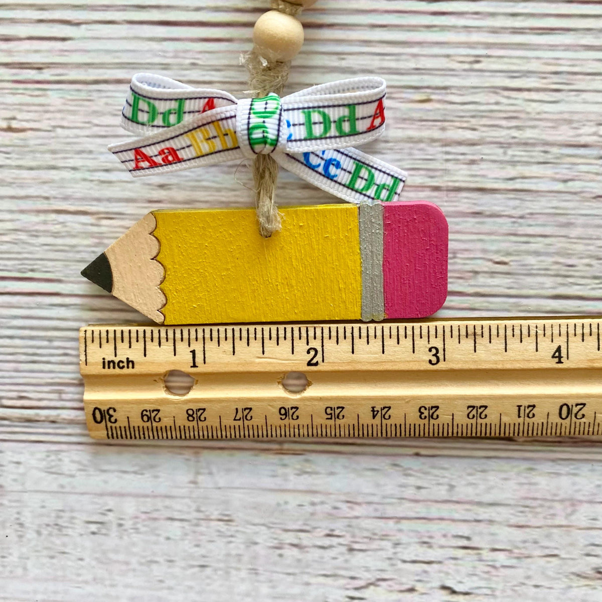 A wooden pencil-shaped ornament with a yellow body, pink eraser, black tip, and a white ribbon bow printed with colorful letters, shown above a wooden ruler on a whitewashed wood background.