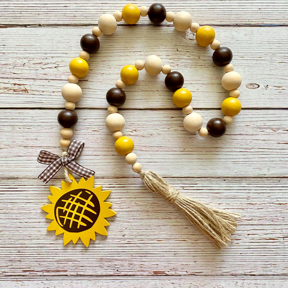 A wooden bead garland with yellow, dark brown, and natural beige beads, featuring a yellow sunflower charm with a gingham ribbon bow and a beige jute tassel, displayed on a whitewashed wooden background.
