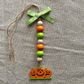 A handmade Halloween hanging ornament featuring a green patterned ribbon bow, a vertical string of orange, green, and cream wooden beads, and a carved jack-o'-lantern pumpkin charm at the bottom, displayed on a textured gray linen background.