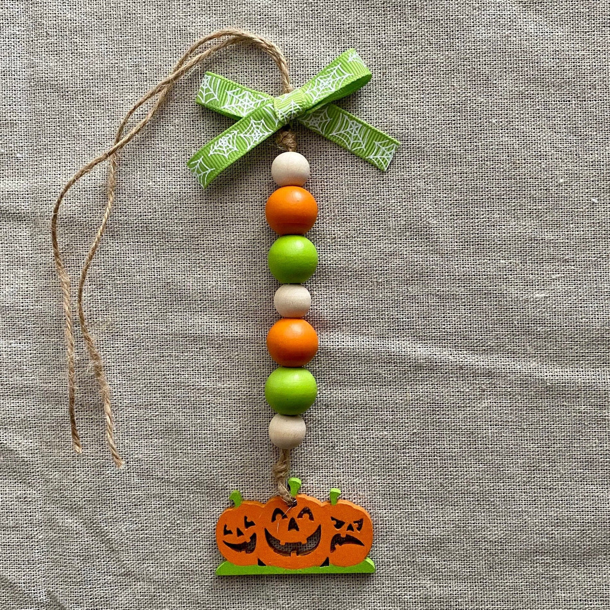 A handmade Halloween hanging ornament featuring a green patterned ribbon bow, a vertical string of orange, green, and cream wooden beads, and a carved jack-o'-lantern pumpkin charm at the bottom, displayed on a textured gray linen background.