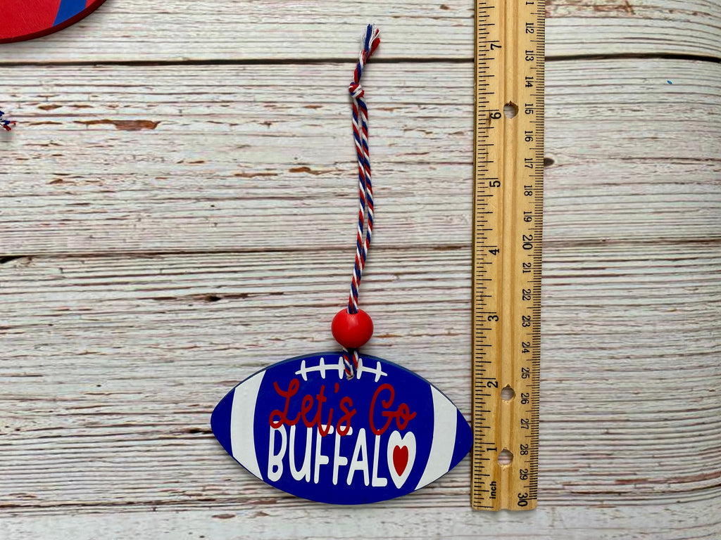 A wooden football-shaped ornament with 'Let's Go BUFFALO' text in red and white, hanging from a red and white striped string next to a wooden ruler on a whitewashed wood background.