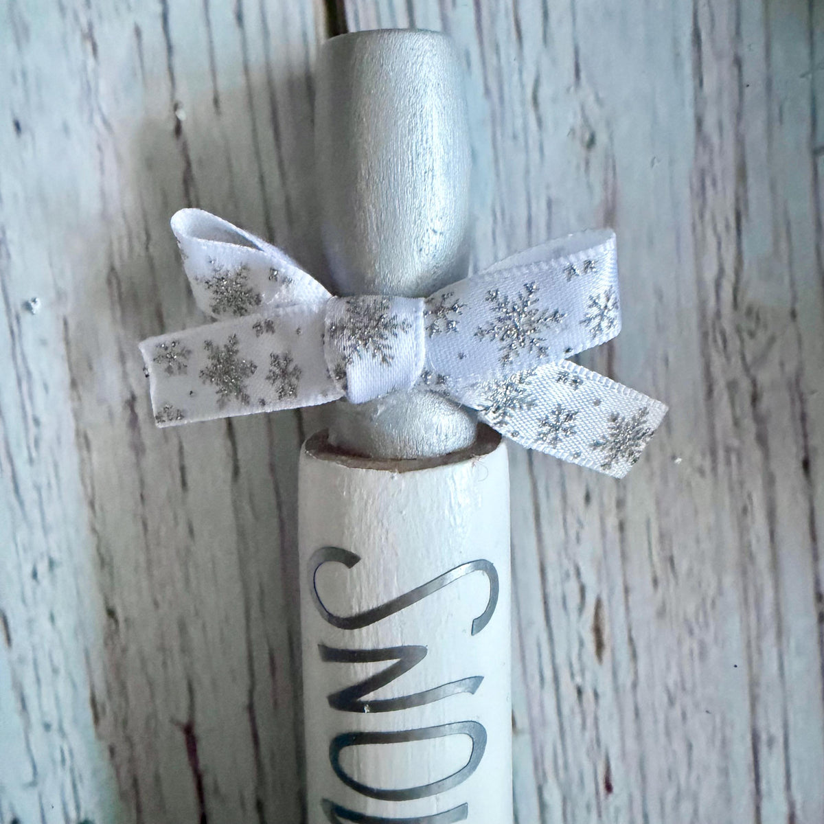 A close-up of a decorative holiday ornament shaped like a rolled scroll or letter, with a silver top, white scroll body, silver lettering, and a white ribbon bow printed with silver snowflakes, set against a distressed whitewashed wood background.