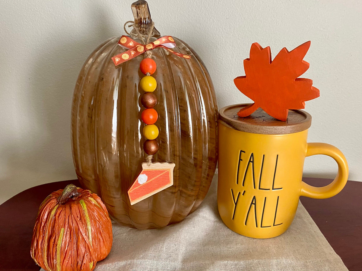 Autumn-themed tabletop decor featuring a large ribbed glass pumpkin with a decorative bow, bead garland, and pie slice ornament, next to a yellow ceramic mug with 'FALL Y'ALL' text and a wooden leaf on top, plus a small striped orange pumpkin on a neutral fabric surface.