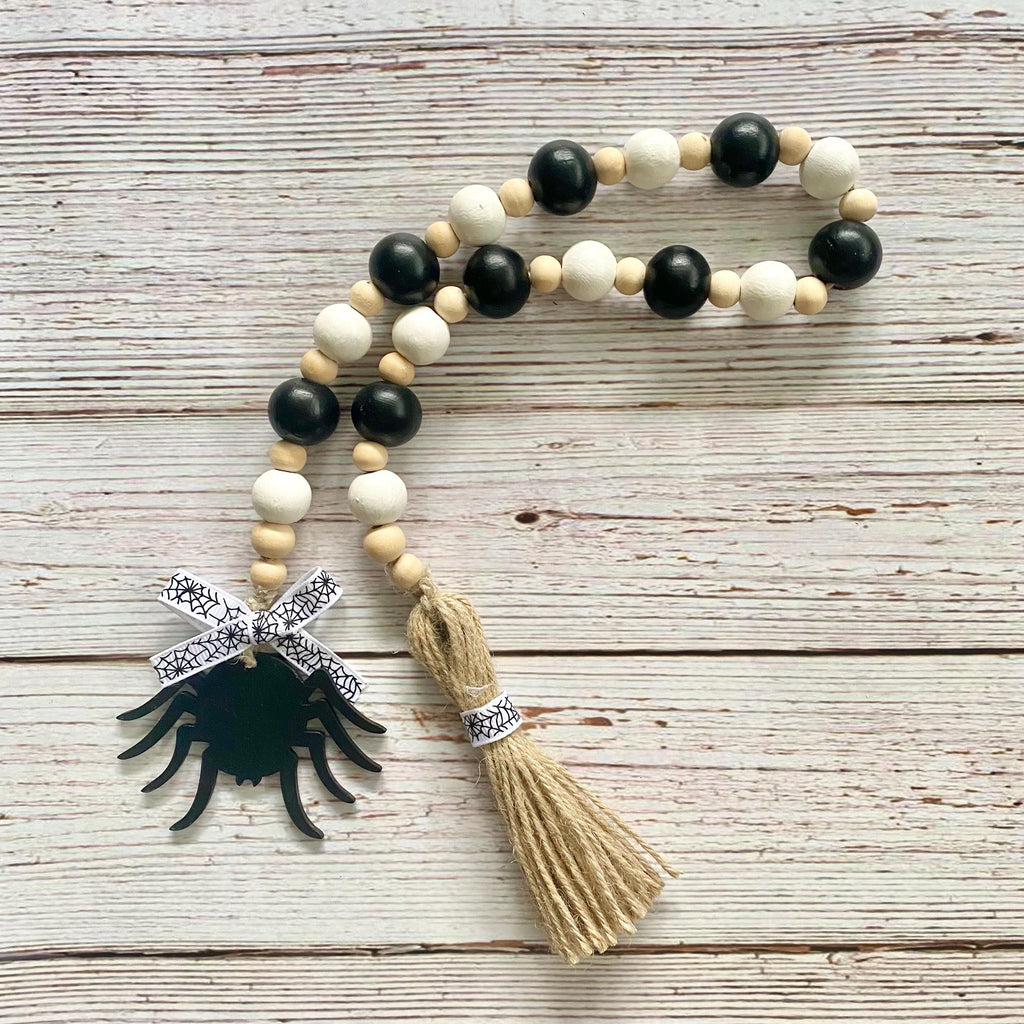 A beaded necklace with alternating black and cream wooden beads, a black spider charm, a cream tassel, and a black-and-white patterned ribbon, displayed on a rustic whitewashed wood background.
