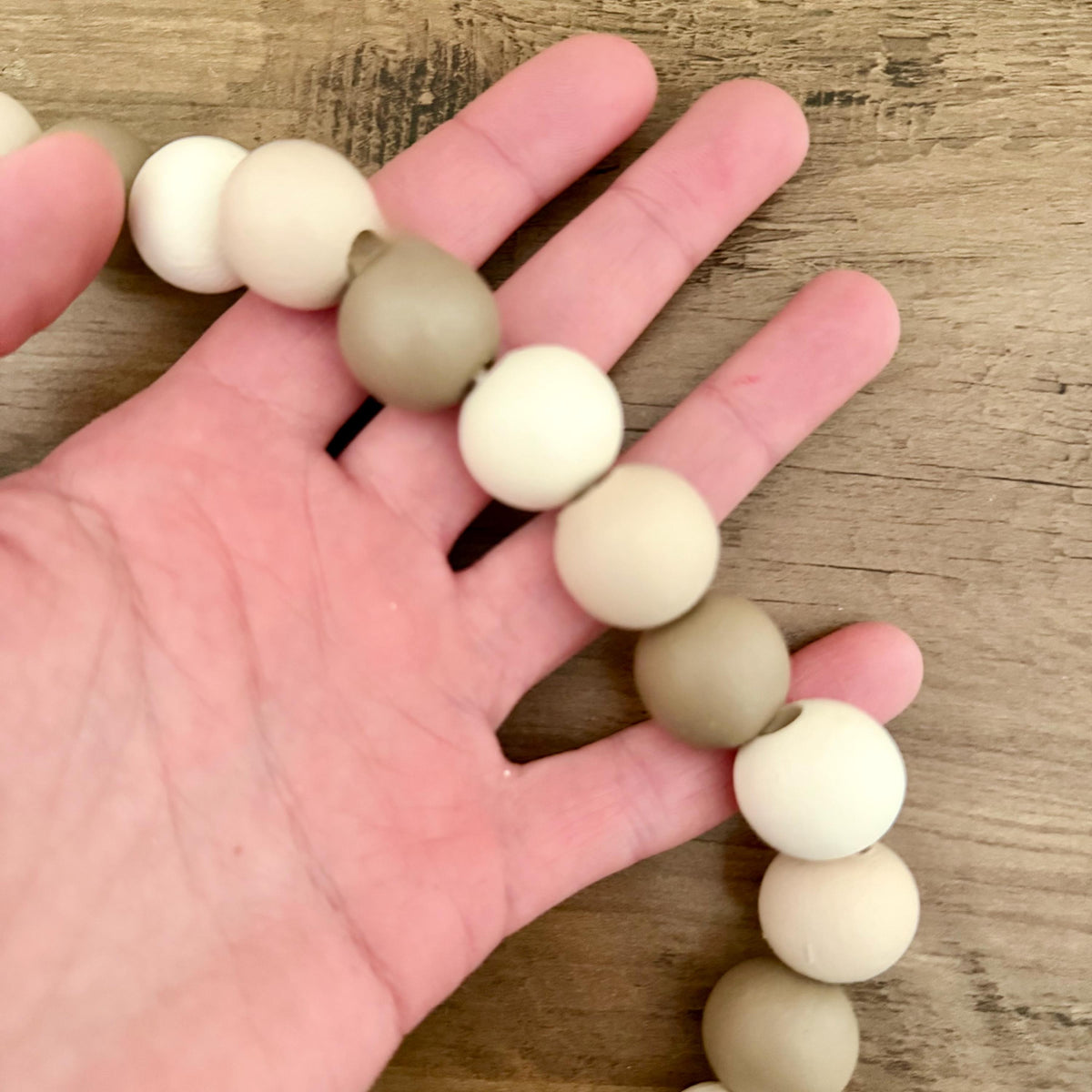 A close-up of a hand holding a beaded string of matte wooden beads in cream and light taupe, resting on a rustic wood surface.
