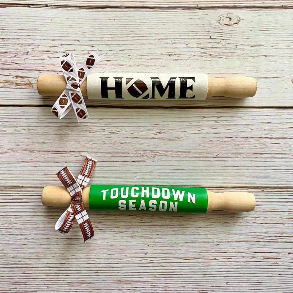 Two wooden rolling pins displayed on a whitewashed wood background. The top rolling pin is light wood with a cream band reading 'HOME' in black, decorated with a white ribbon printed with football patterns.