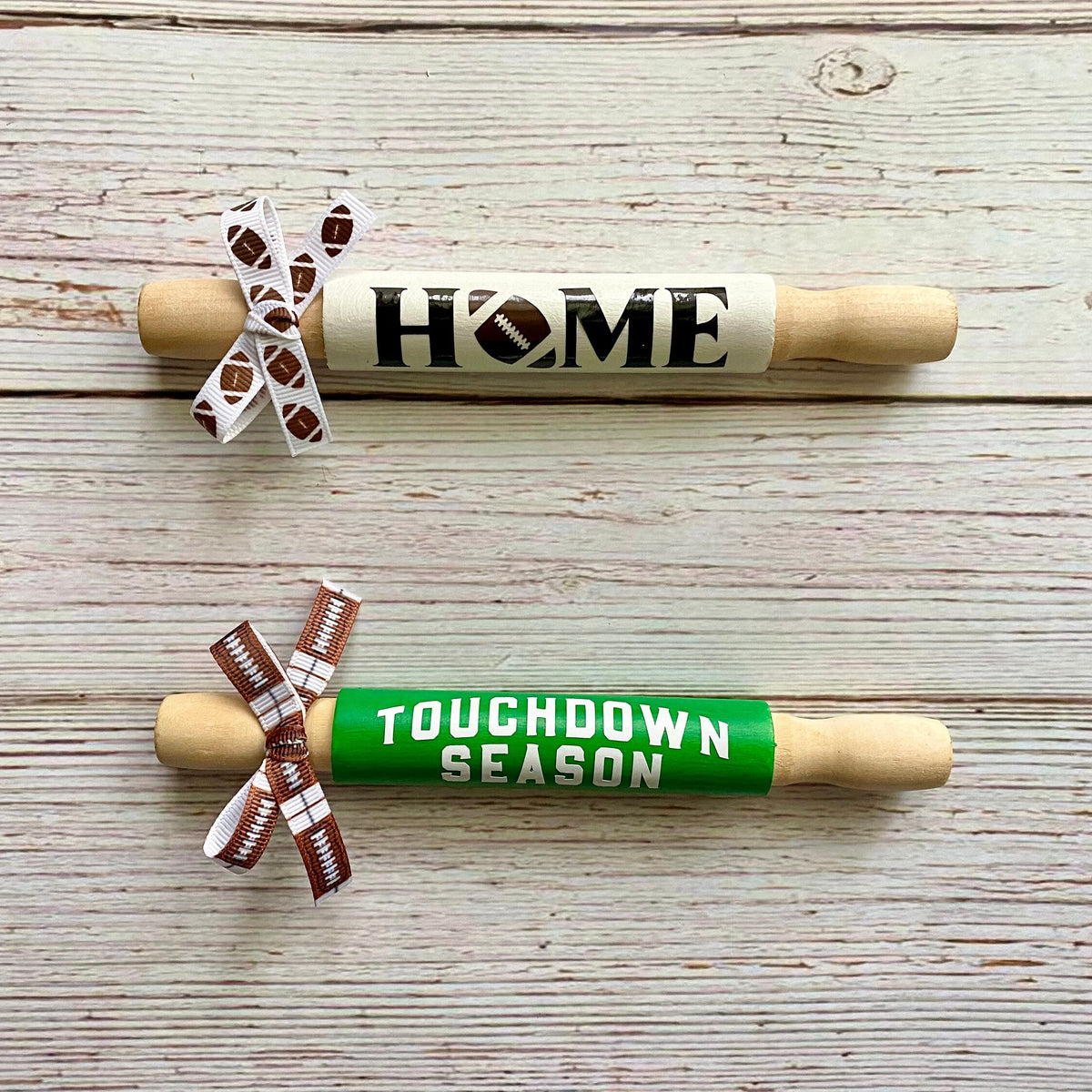 Two wooden rolling pins displayed on a whitewashed wood background. The top rolling pin is light wood with a cream band reading 'HOME' in black, decorated with a white ribbon printed with football patterns.