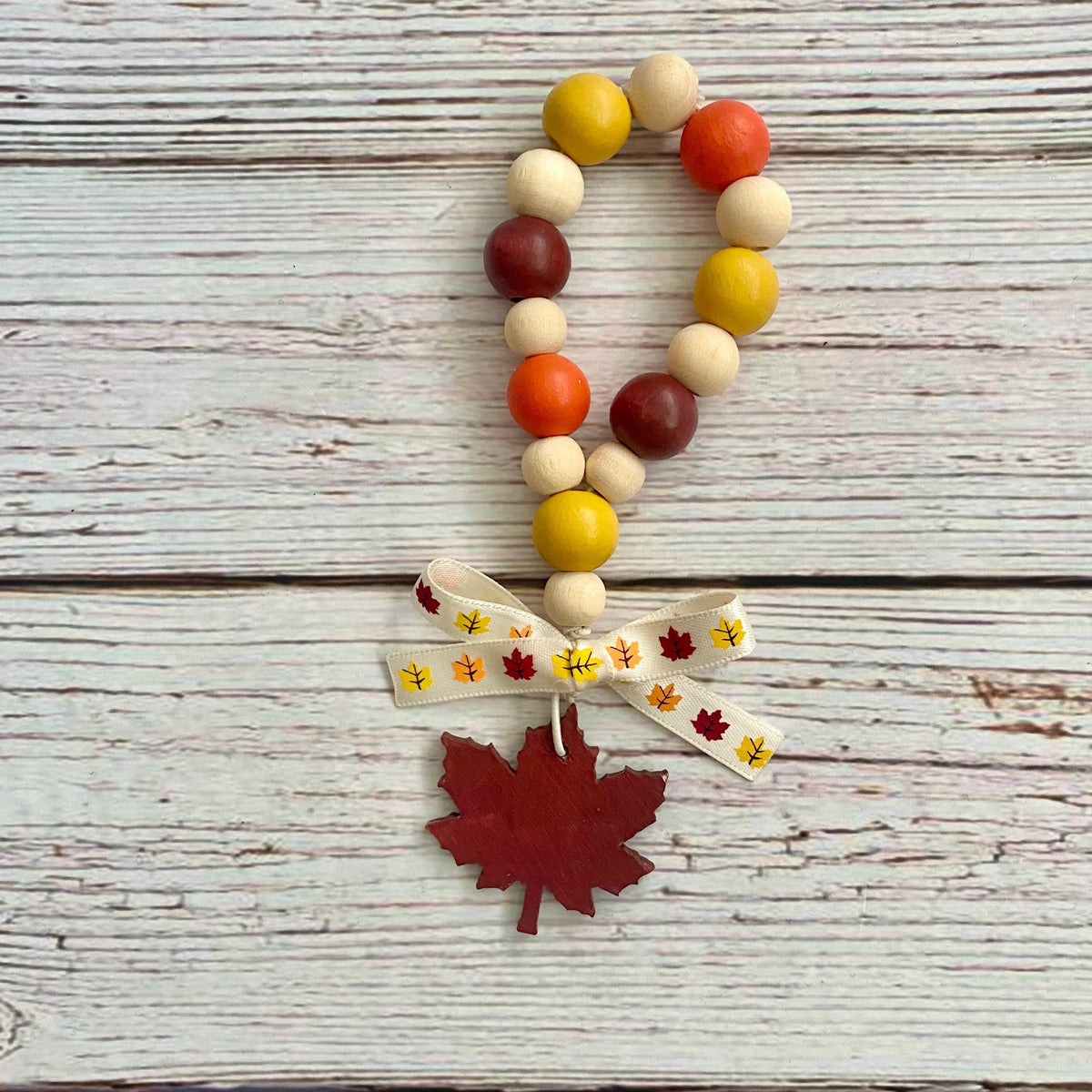 Autumn-themed wooden bead garland with orange, yellow, and dark red round beads, a cream ribbon bow printed with maple leaves, and a red wooden maple leaf charm, displayed on a white-washed wood background.