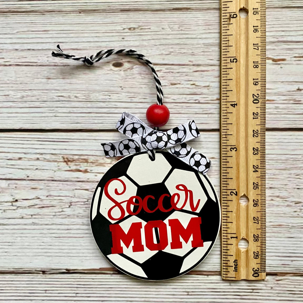 A round soccer-themed ornament shaped like a soccer ball with the words 'Soccer MOM' in red script and block lettering, hanging from a black-and-white striped string with a red bead and black-and-white patterned ribbon. The ornament is photographed on a whitewashed wood surface next to a wooden ruler for scale.