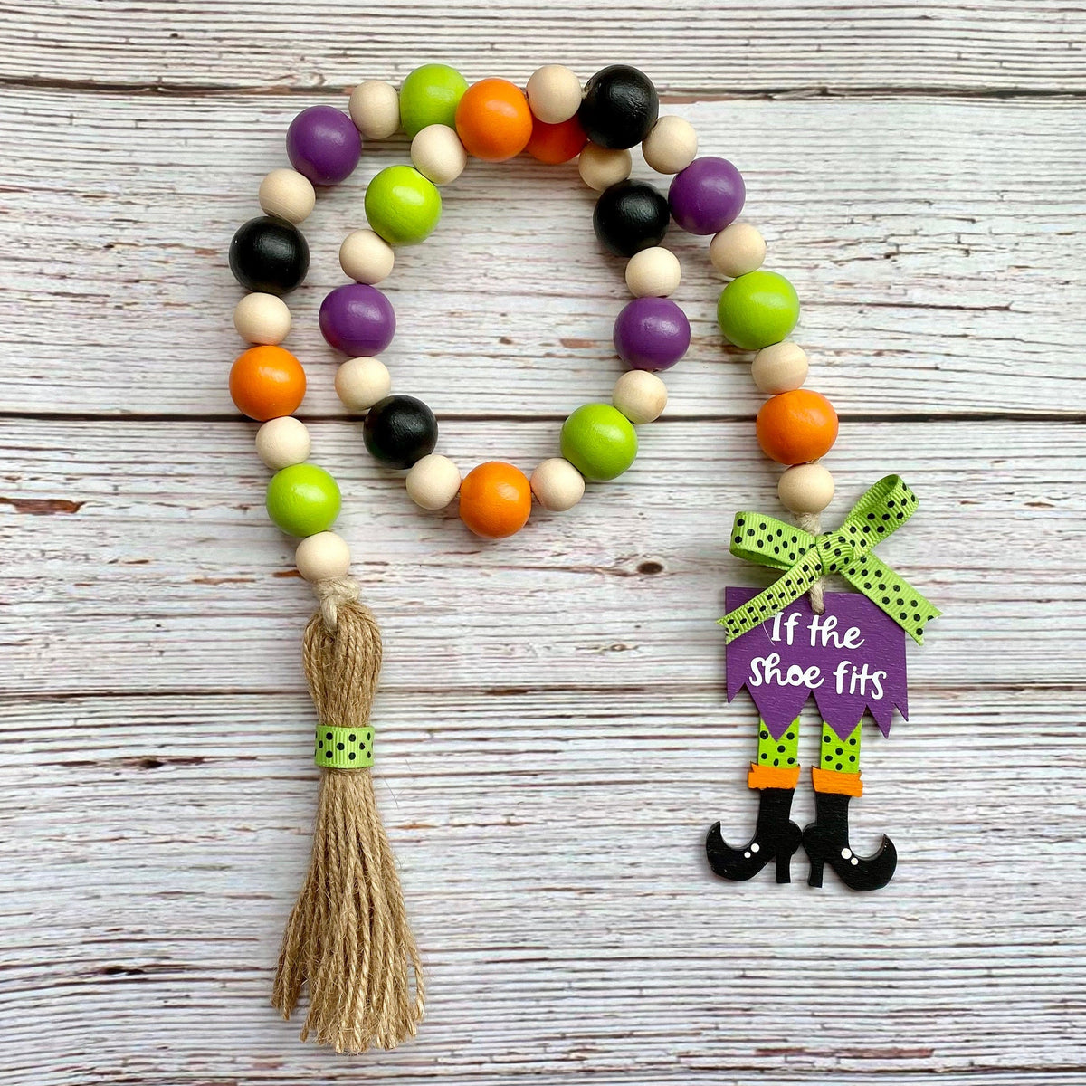 A wooden bead garland with orange, green, purple, black, and natural wood beads, featuring a tassel and a witch character charm that reads 'If the shoe fits', displayed on a rustic white wood background.
