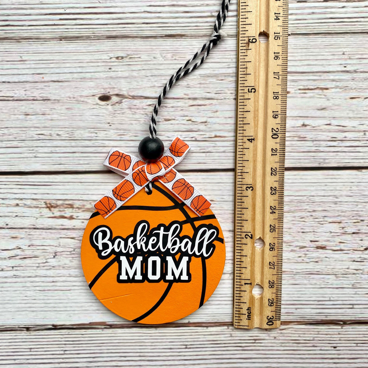 A round basketball-themed ornament or plaque with the words 'Basketball MOM' in white script and block lettering, decorated with small basketball illustrations at the top and a black-and-white twisted string. It is photographed on a whitewashed wood surface next to a wooden ruler for scale.
