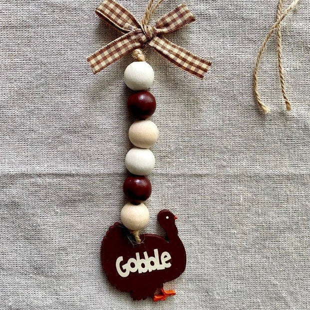 A rustic fall-themed wooden ornament featuring a turkey-shaped charm with the word 'Gobble' in white, strung with cream and dark red wooden beads, and finished with a brown gingham ribbon bow at the top, displayed on a textured gray linen background.