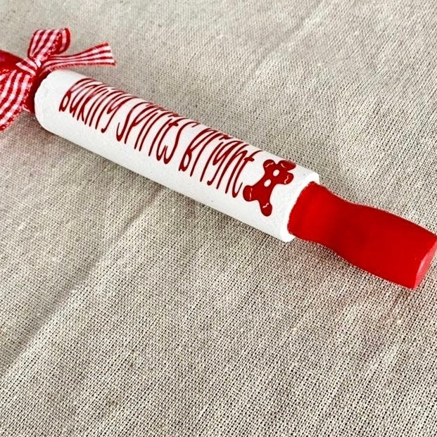 A white rolling pin with red handles and red decorative lettering that reads 'Baking Spices Kitchen,' plus a red-and-white gingham ribbon bow, placed on a beige fabric background.