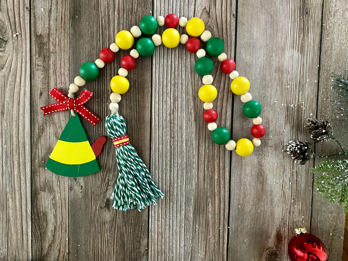 A Christmas-themed wooden bead garland with red, green, yellow, and cream beads, featuring a green and yellow elf hat ornament with a red bow and a green and white striped tassel, displayed on a rustic wooden background.