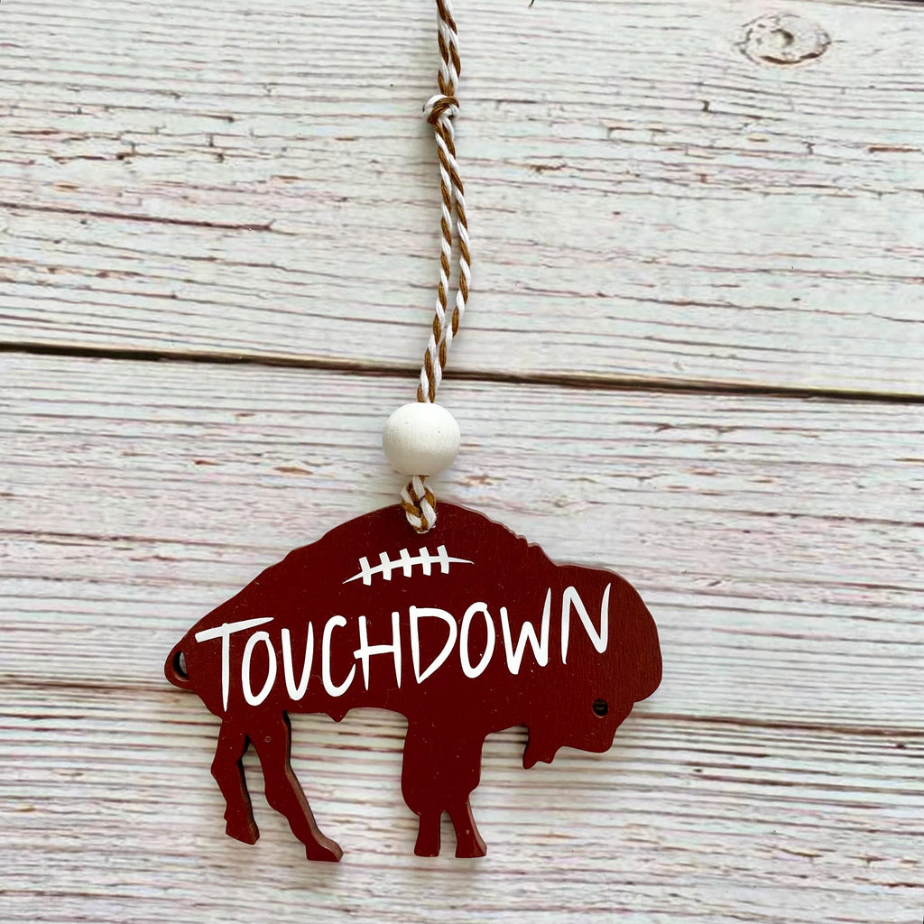 A buffalo-shaped wooden ornament in dark red with the word 'TOUCHDOWN' in white, hanging from a twisted string with a white bead, set against a whitewashed wood plank background.