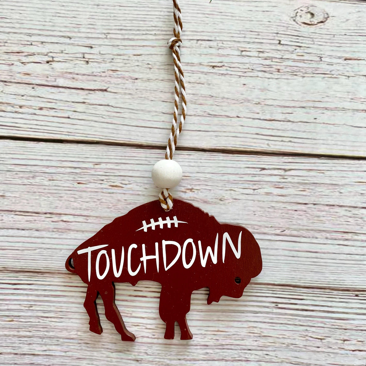 A buffalo-shaped wooden ornament in dark red with the word 'TOUCHDOWN' in white, hanging from a twisted string with a white bead, set against a whitewashed wood plank background.