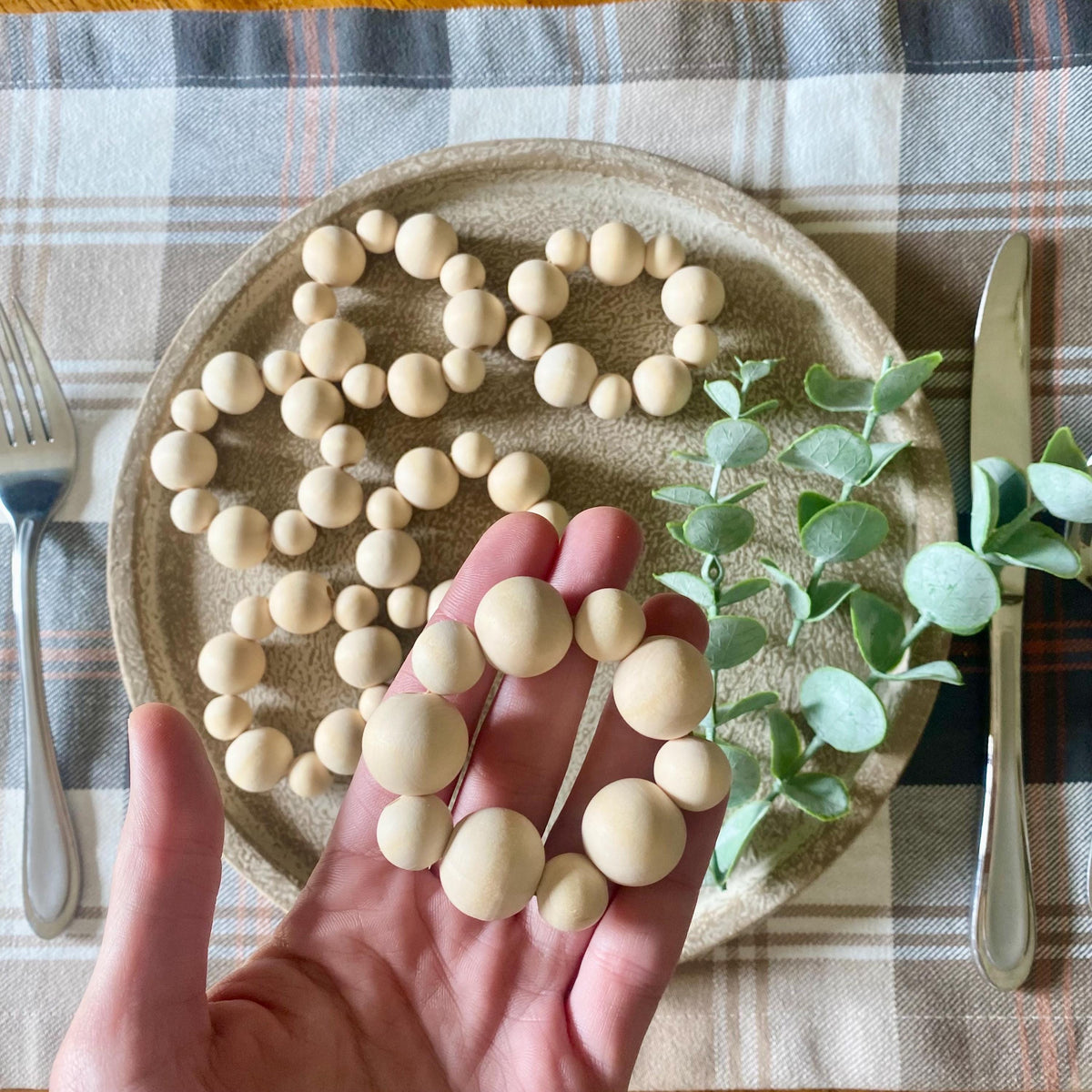 A hand holds a ring made of round, matte cream-colored beads, with more matching beads scattered on a ceramic plate and green eucalyptus stems beside it on a plaid tablecloth.