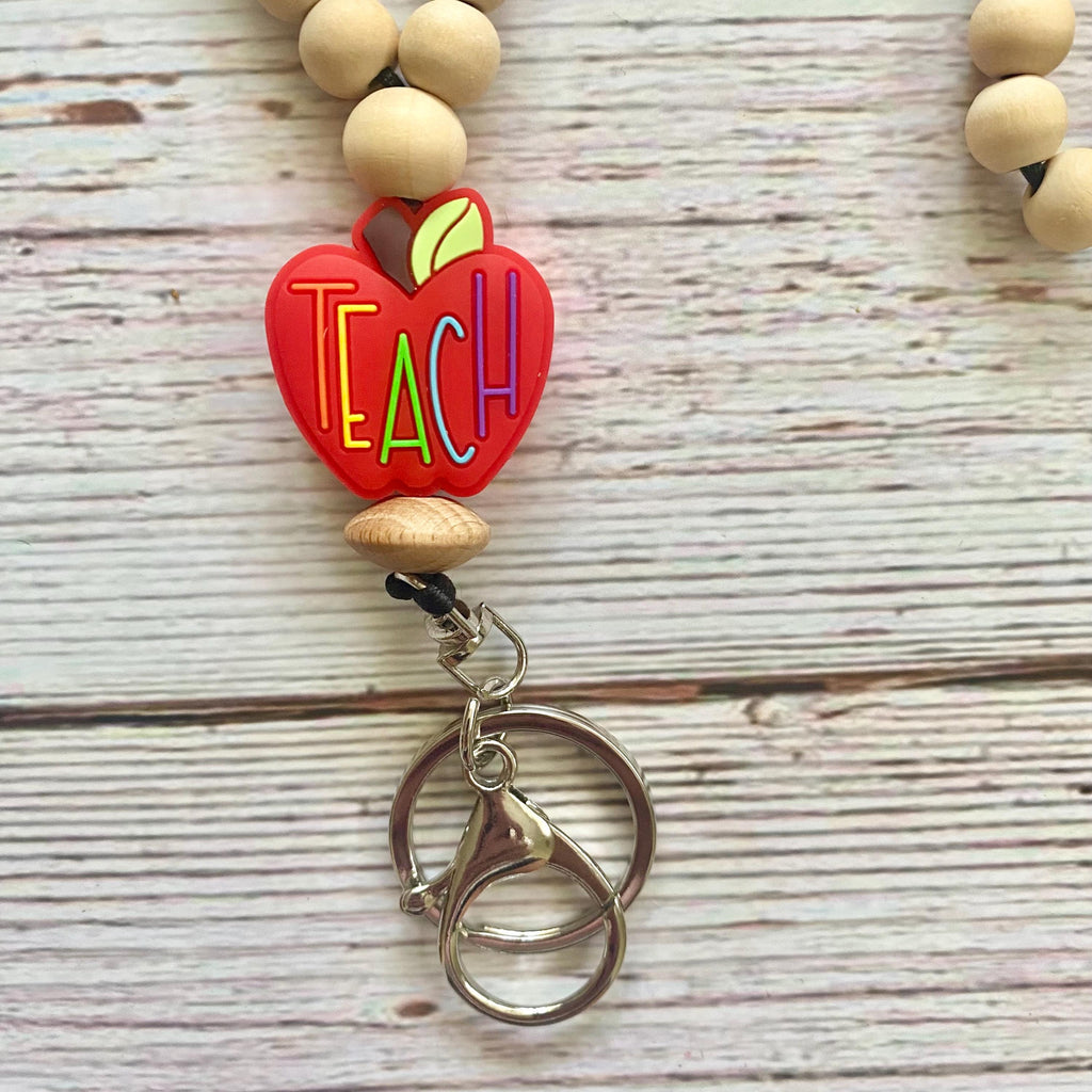 A beaded keychain featuring a red apple charm with the word 'TEACH' in colorful letters, suspended from natural wood beads on a whitewashed wooden background.