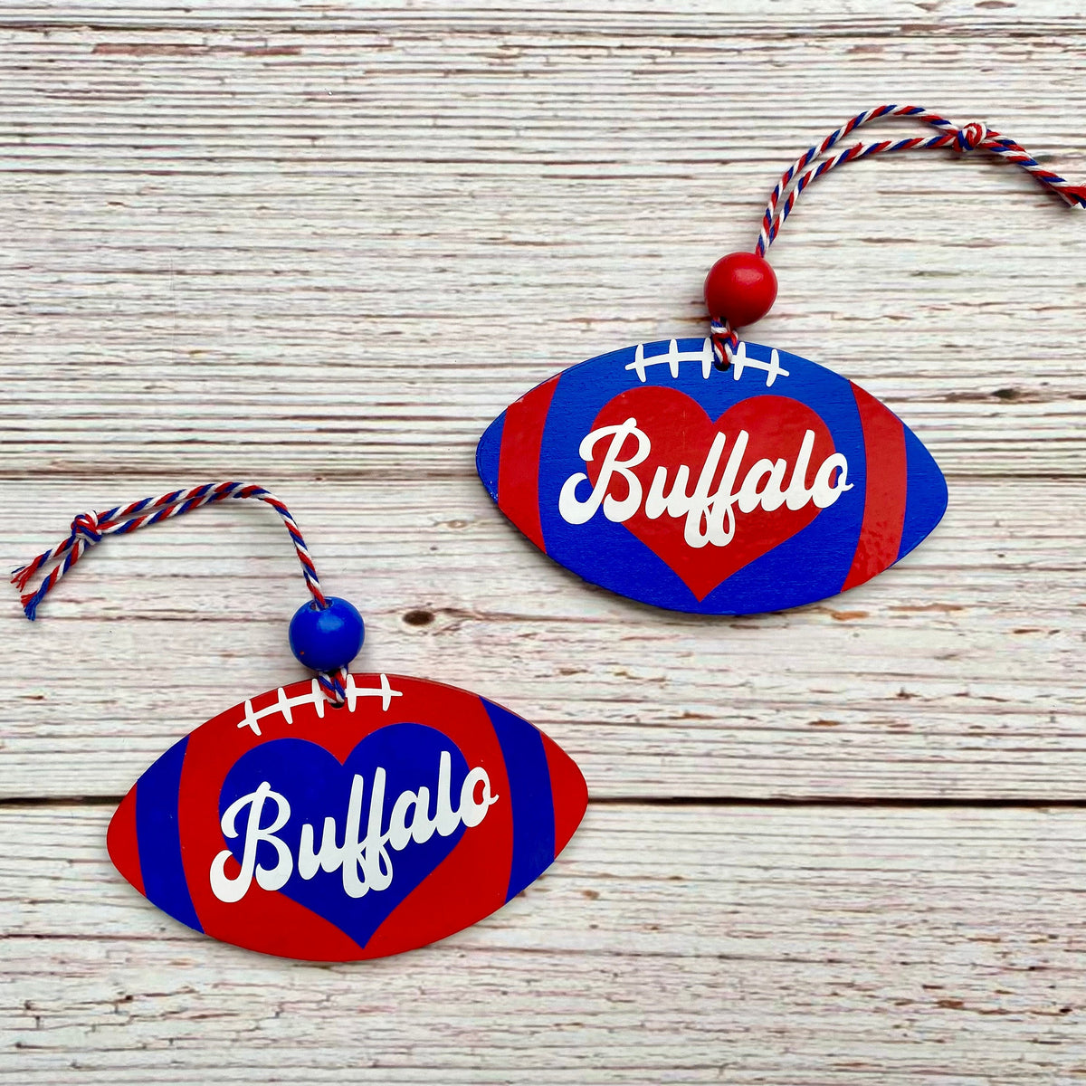 Two Buffalo football-shaped ornaments on a whitewashed wooden surface. Each ornament has a red and blue striped body, a red heart in the center with the word 'Buffalo' in white script, and a hanging loop made of red, white, and blue cord with a red bead.
