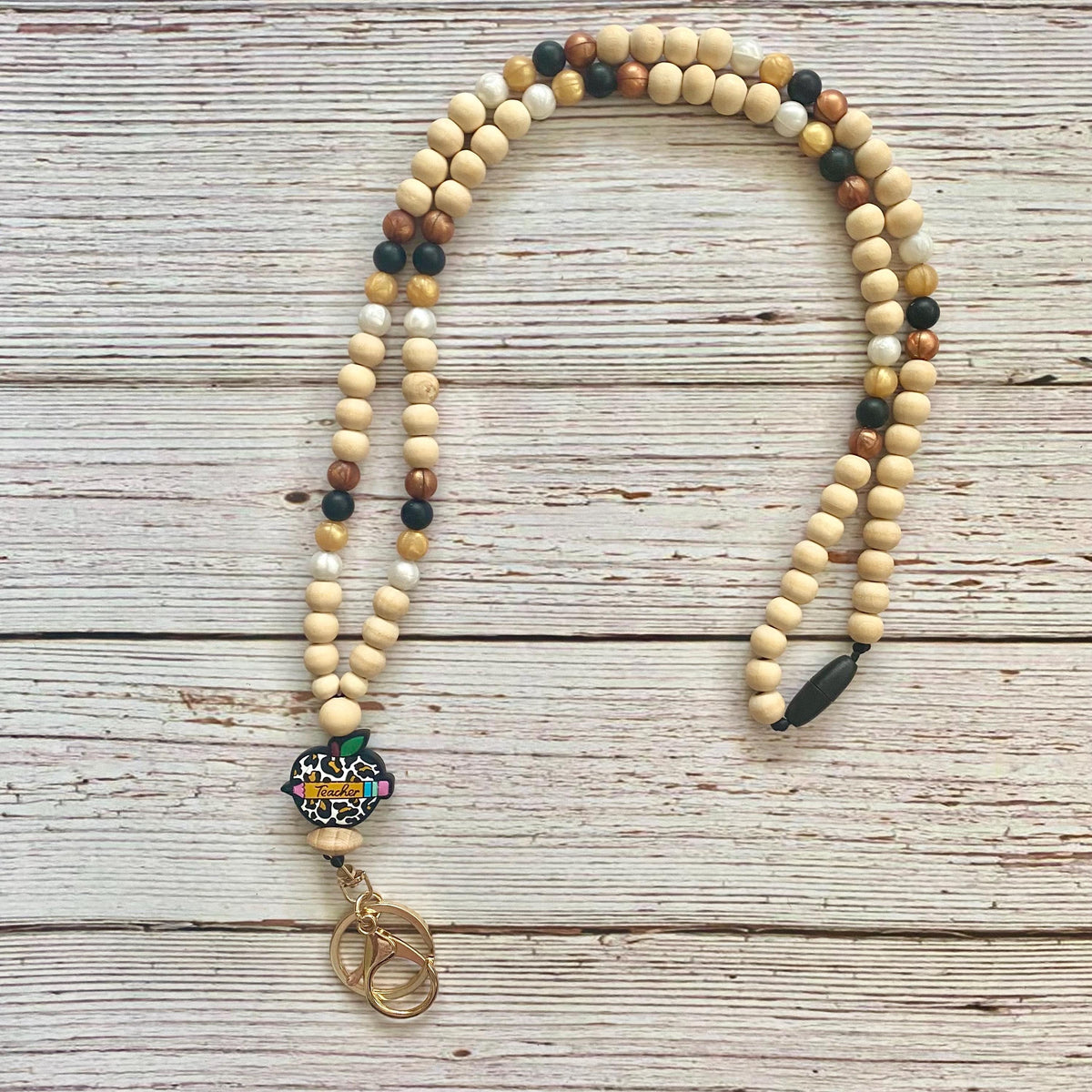 A beaded necklace with natural wood beads in cream, tan, brown, and black, featuring a colorful floral charm and a gold-toned metal clasp, laid flat on a whitewashed wooden surface.