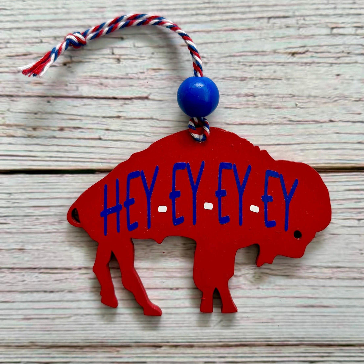 A red buffalo silhouette ornament with blue 'HEY EY EY EY' lettering, hanging from a red, white, and blue string with a blue bead, set against a rustic white-washed wood background.