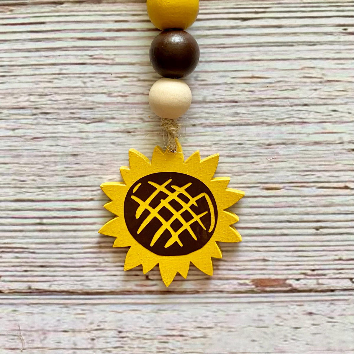 A wooden sunflower ornament with a yellow painted sunflower head, a dark brown center, and a string of wooden beads in yellow, brown, and cream, displayed on a whitewashed wooden surface.