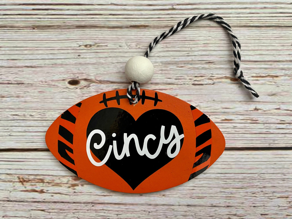 A football-shaped ornament with orange and black tiger-stripe patterns, a black heart in the center, white script text reading 'Cincy,' and a black-and-white hanging cord on a whitewashed wood background.