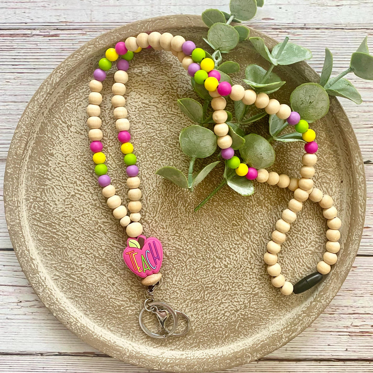 A beaded necklace with natural wood beads and bright pink, yellow, and purple accents, featuring a pink heart charm with a letter 'I' and a silver double-heart pendant, displayed on a textured beige plate with green leaves.