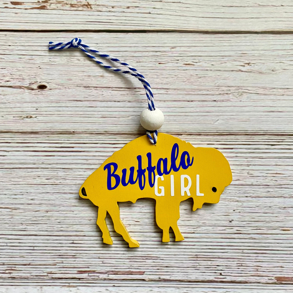 A yellow silhouette ornament shaped like a buffalo with the words 'Buffalo GIRL' printed on it, hanging from a blue-and-white string with a white bead, set against a rustic whitewashed wood background.
