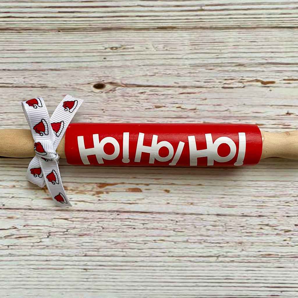 A wooden rolling pin wrapped in a bright red paper sleeve printed with 'Ho! Ho! Ho!' in white, tied with a white ribbon featuring red Santa hats, set on a rustic white-washed wood background.