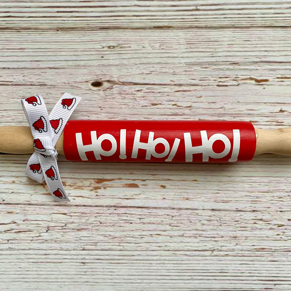 A wooden rolling pin wrapped in a bright red paper sleeve printed with 'Ho! Ho! Ho!' in white, tied with a white ribbon featuring red Santa hats, set on a rustic white-washed wood background.