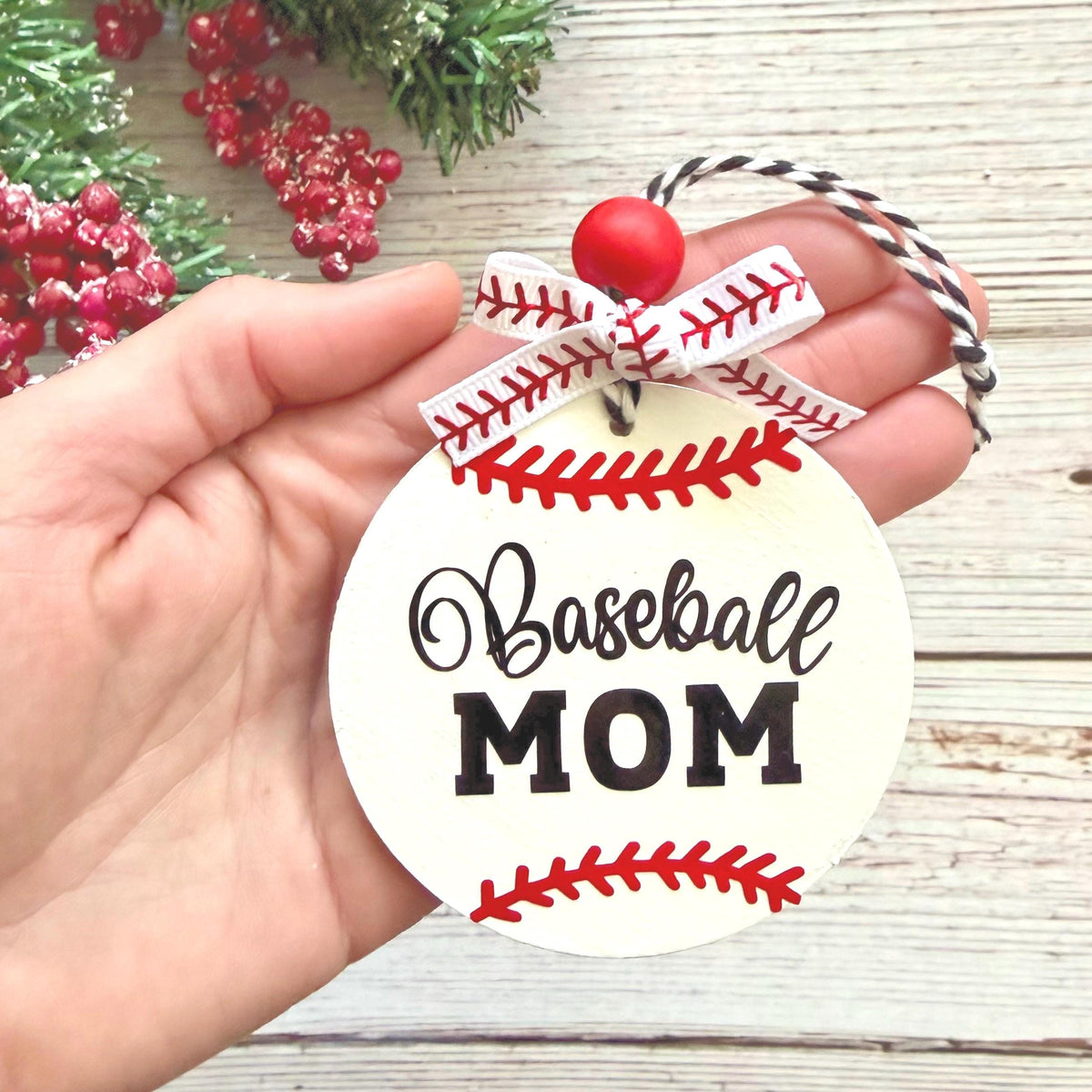 A hand holds a round white ornament designed like a baseball, with red stitching details, a red bead, a white and red ribbon bow, and the words 'Baseball MOM' in black script and block lettering. The background shows a light wood surface with some greenery and red berries.