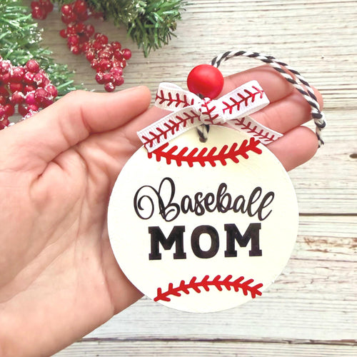 A hand holds a round white ornament designed like a baseball, with red stitching details, a red bead, a white and red ribbon bow, and the words 'Baseball MOM' in black script and block lettering. The background shows a light wood surface with some greenery and red berries.