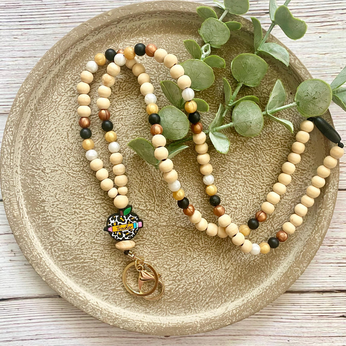 A beaded necklace with light wood beads, small brown and black accent beads, and a colorful leopard-print charm with a gold-tone key ring, displayed on a textured beige ceramic plate with green eucalyptus leaves.