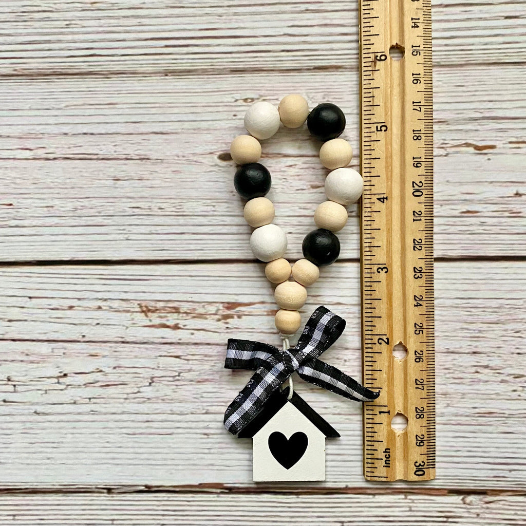 A handmade wooden bead garland ornament with a black and white gingham ribbon bow and a small white house charm with a black heart, displayed next to a wooden ruler on a whitewashed wood plank background.
