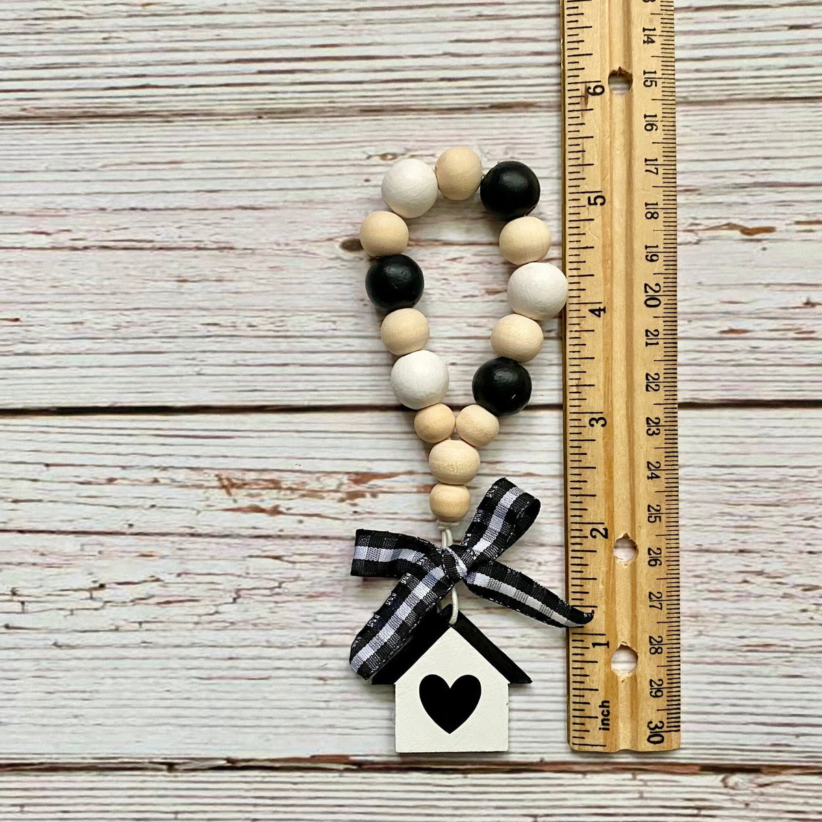 A handmade wooden bead garland ornament with a black and white gingham ribbon bow and a small white house charm with a black heart, displayed next to a wooden ruler on a whitewashed wood plank background.