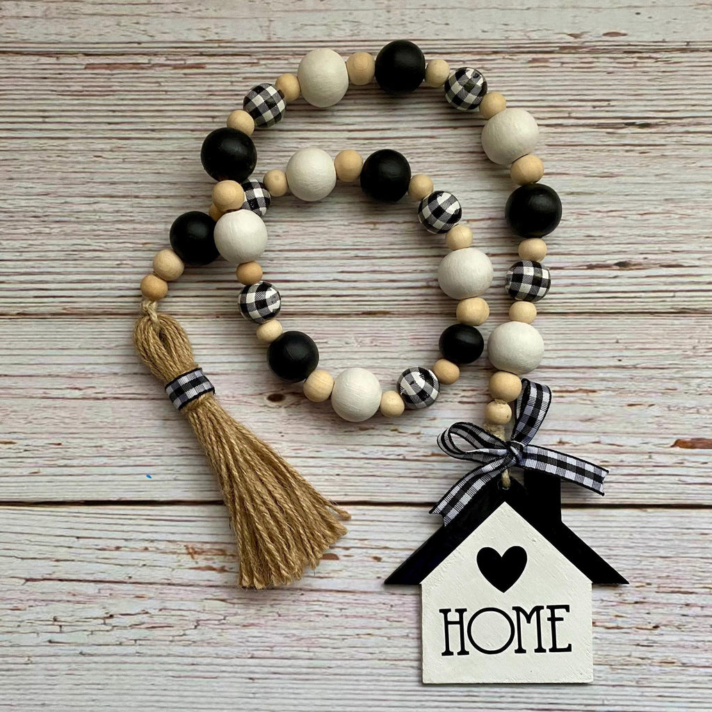 A rustic wooden bead garland with black, white, and buffalo plaid accents, a jute tassel, and a hanging house-shaped ornament that says HOME, displayed on a whitewashed wood background.
