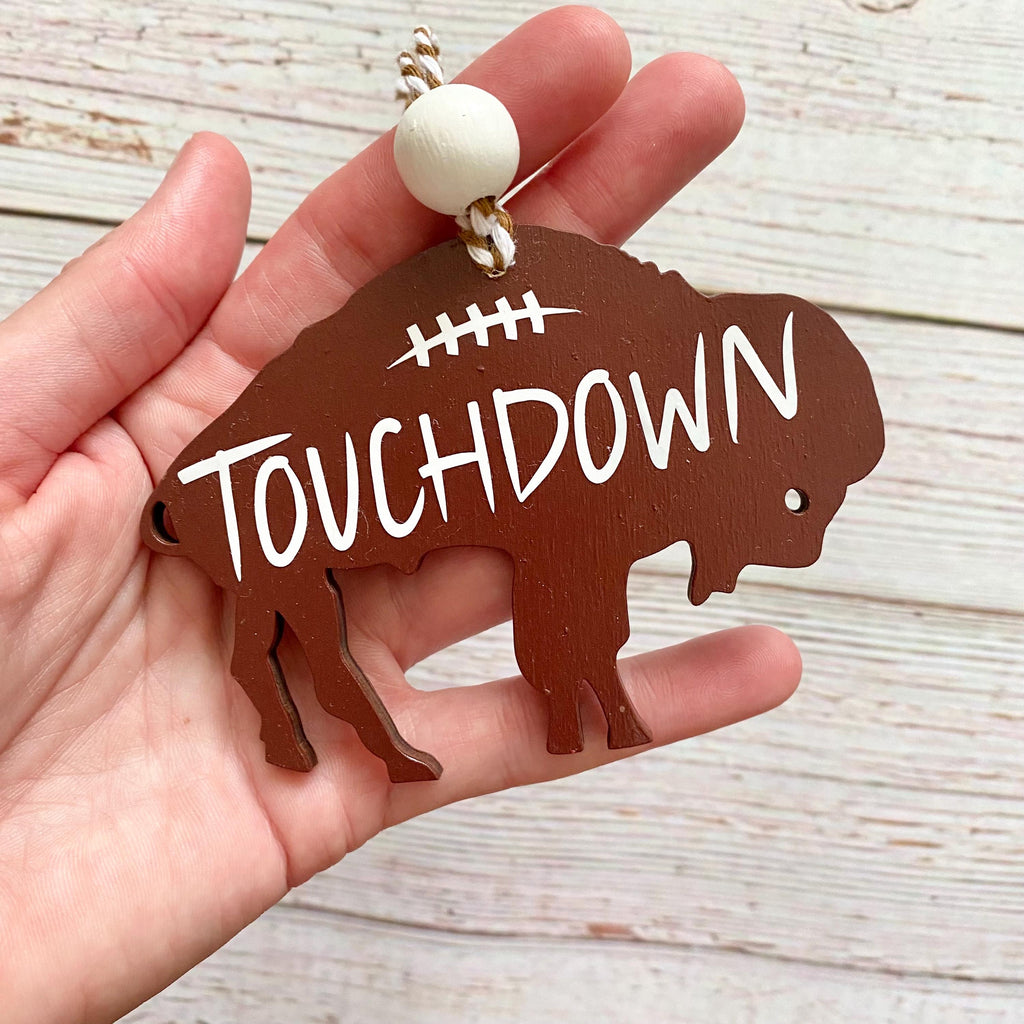 A hand holding a brown wooden bison-shaped ornament with the word 'TOUCHDOWN' in white lettering and a white bead at the top, set against a light wood plank background.
