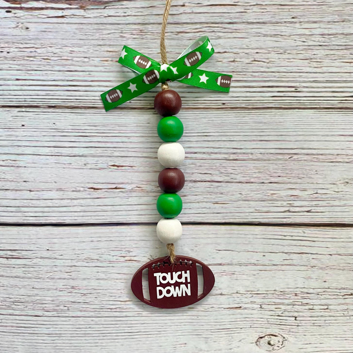 A decorative hanging ornament with a green ribbon bow printed with footballs and stars, a vertical string of green, white, and brown beads, and a brown football-shaped tag that reads “TOUCH DOWN,” displayed against a whitewashed wood plank background.