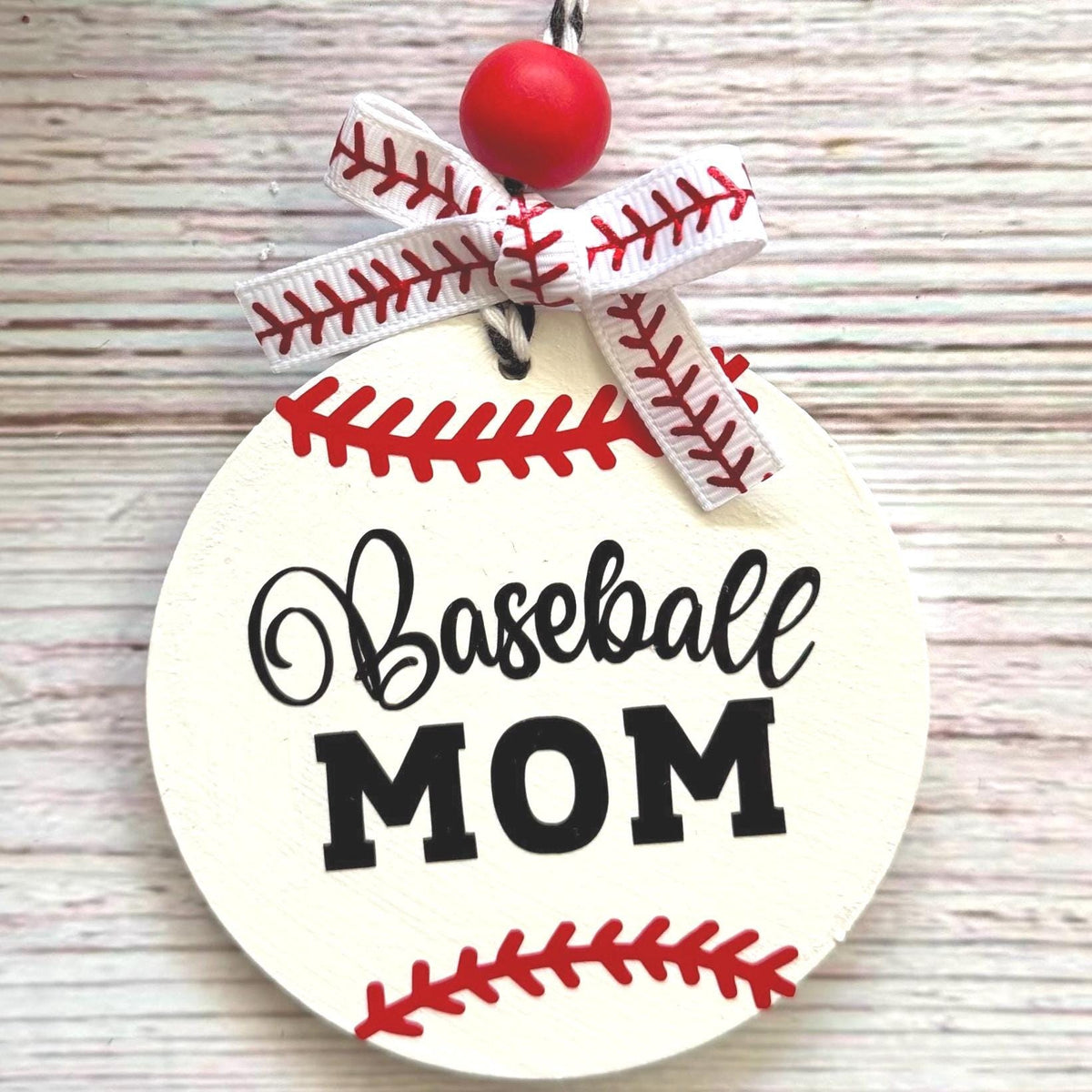 A round wooden ornament designed like a baseball, with the words 'Baseball MOM' in black script and block lettering, red seam details, a white ribbon bow with red stitching, and a red hanging bead, shown on a light wood background.