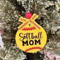 A round yellow softball ornament hanging on a frosted Christmas tree. The ornament features the words 'Softball MOM' in black script and block lettering, decorated with red baseball-style stitching details and a yellow ribbon bow at the top.