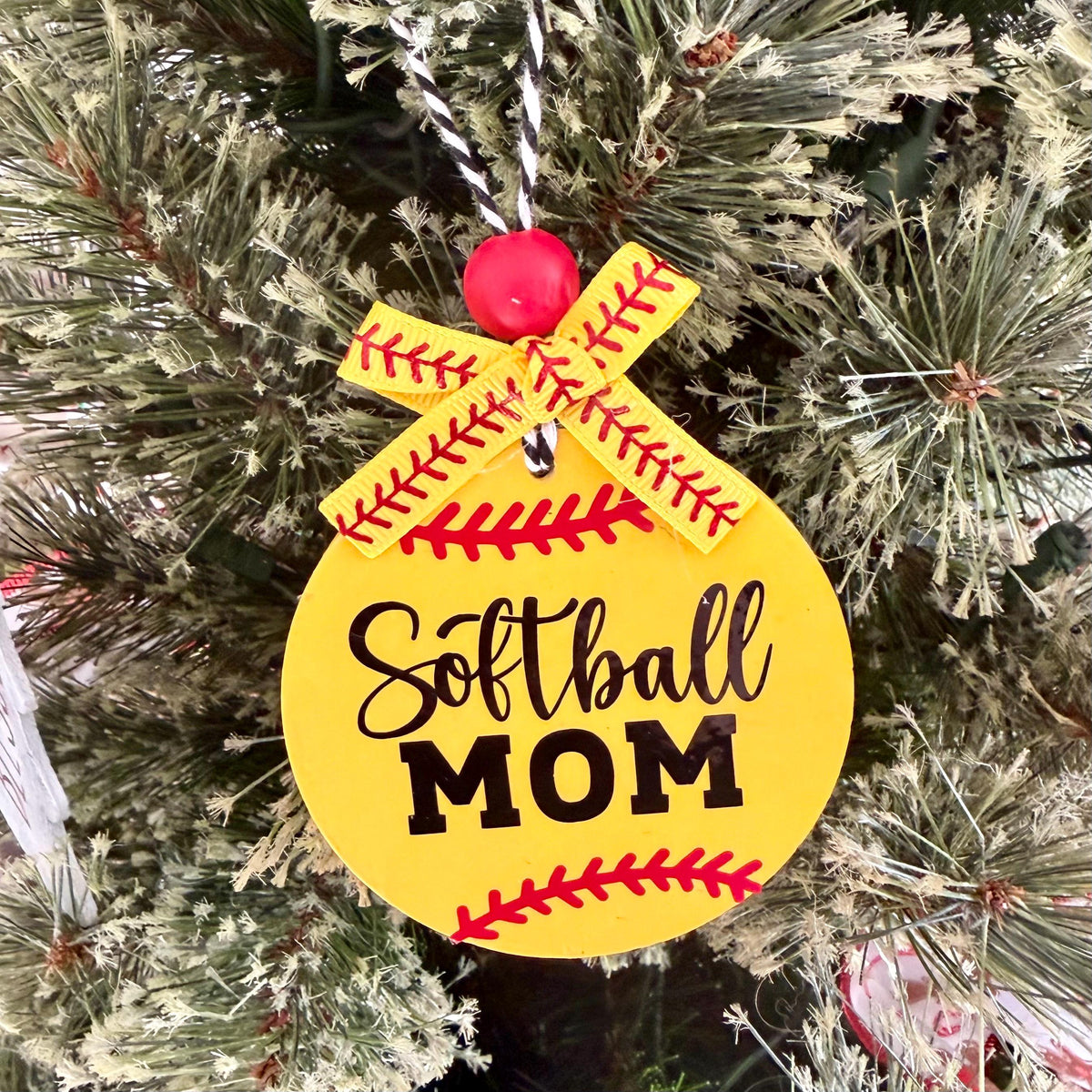 A round yellow softball ornament hanging on a frosted Christmas tree. The ornament features the words 'Softball MOM' in black script and block lettering, decorated with red baseball-style stitching details and a yellow ribbon bow at the top.