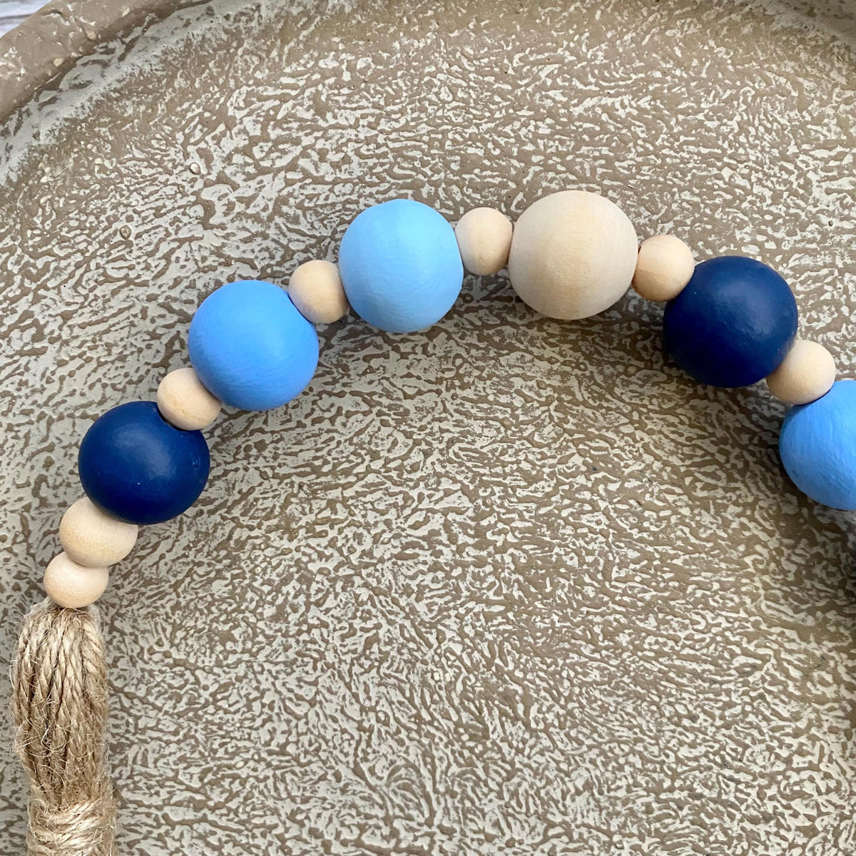 A beaded garland made of alternating light blue, dark navy, and natural beige wooden beads, with a knotted jute tassel, displayed on a textured beige surface.