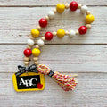 A beaded garland with red, yellow, and white wooden beads, featuring a black chalkboard charm with 'ABC' and a small red apple, plus a red-and-white striped paper tassel, displayed on a light wood background.