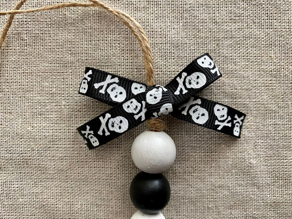 A decorative hanging ornament with a black ribbon bow printed with white skulls and crossbones, a white wooden bead, and a black wooden bead, shown against a beige linen background.