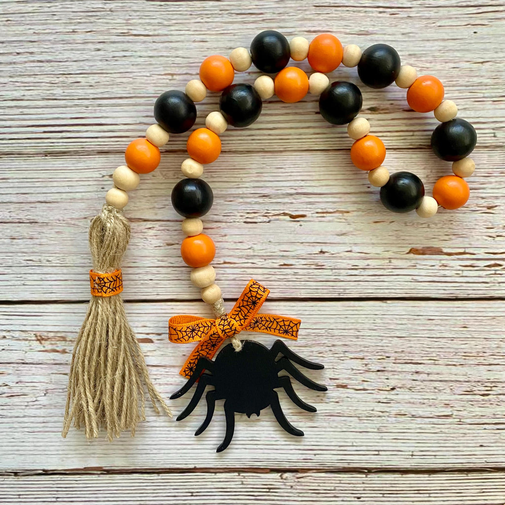 Halloween-themed wooden bead garland with black and orange beads, a beige tassel wrapped in orange patterned ribbon, and a black spider decoration tied with a matching orange bow, displayed on a rustic white wood background.