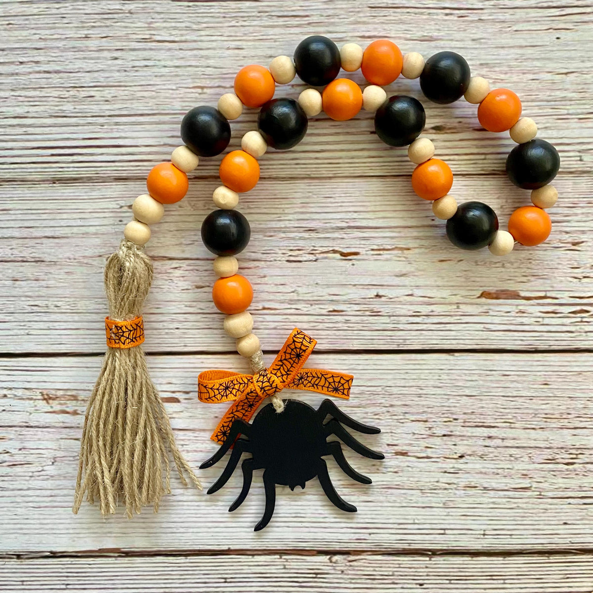 Halloween-themed wooden bead garland with black and orange beads, a beige tassel wrapped in orange patterned ribbon, and a black spider decoration tied with a matching orange bow, displayed on a rustic white wood background.