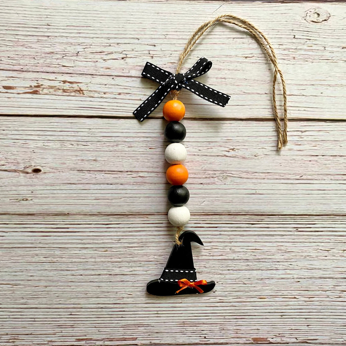 A Halloween-themed wooden bead garland ornament featuring a black witch hat charm, orange and white beads, a black ribbon bow, and a jute hanging string, displayed on a rustic white-painted wooden background.