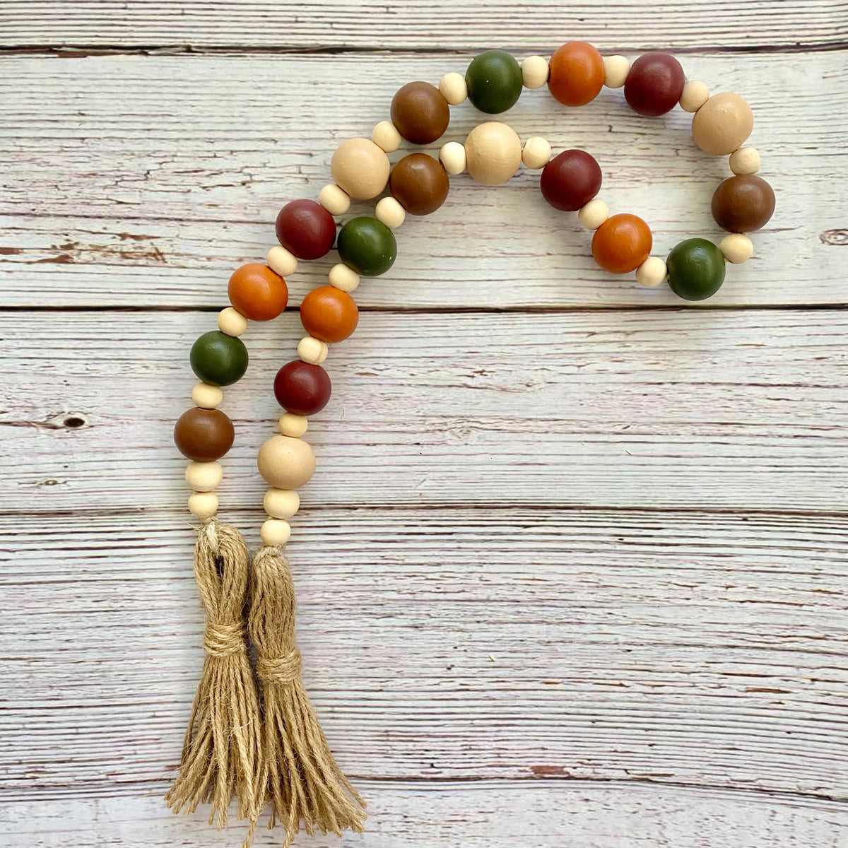 A beaded garland with natural wood beads, round wooden beads in green, orange, and red, and two tan tassels, displayed on a rustic white-washed wooden background.