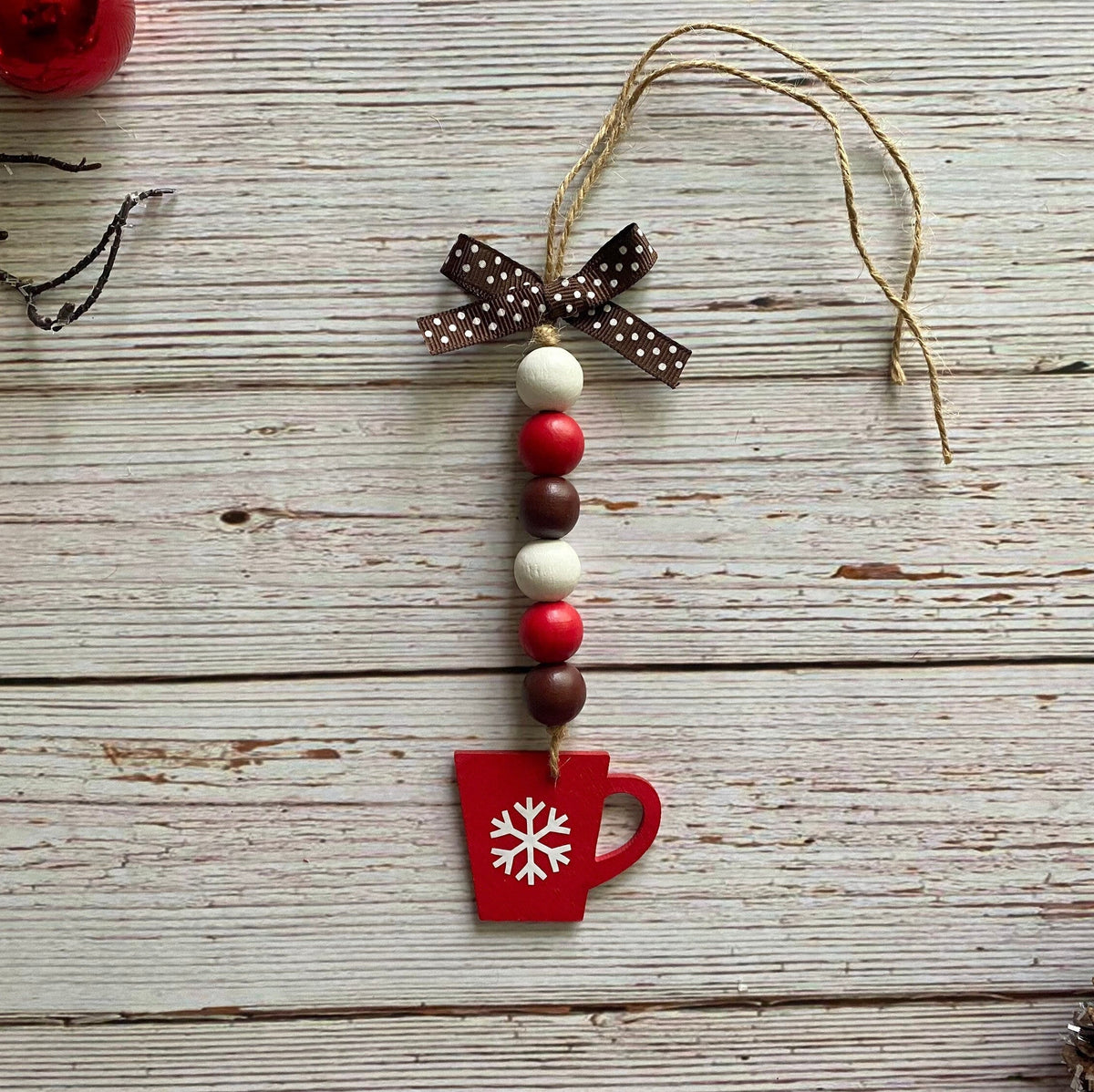 A rustic Christmas ornament shaped like a red mug with a white snowflake, hanging from a brown polka-dot ribbon bow and a string of red, white, and brown beads on a whitewashed wooden background.